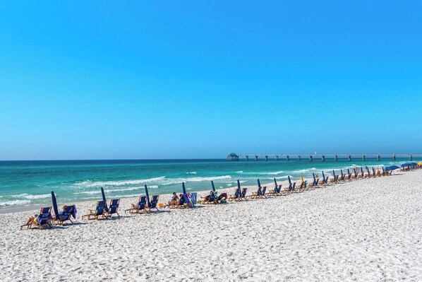 Ein Strand mit Reihen von Liegestühlen und Sonnenschirmen, ein Steg im Hintergrund und ein klarer, blauer Himmel.