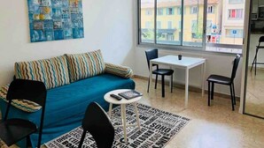 Uma sala de estar moderna com um sofá azul, uma mesinha e duas cadeiras. Há uma janela grande com vista para edifícios.