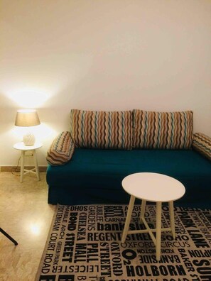 Una habitación con un sofá color turquesa, un cojín estampado, una mesita blanca y una lámpara.