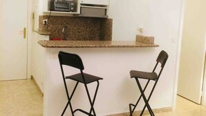Cozinha compacta com micro-ondas, torradeira e pia. Há duas cadeiras dobráveis e um pequeno balcão.