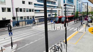 Uma rua urbana moderna com linhas de bonde, um suporte para bicicletas e um prédio com a inscrição 'URGENCES PEDIATRIQUES'.