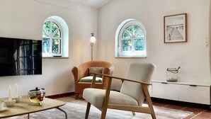 Ein Wohnzimmer mit zwei Rundbogenfenstern, einem Flachbildfernseher, einem Korbstuhl, einem modernen Sessel, einem kleinen Tisch mit Kerzen und einem gerahmten Bild an der Wand.