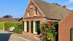 Ein Ziegelhaus mit Satteldach und grünen Fensterläden, ein geparktes Auto und ein Hecken im Vordergrund.