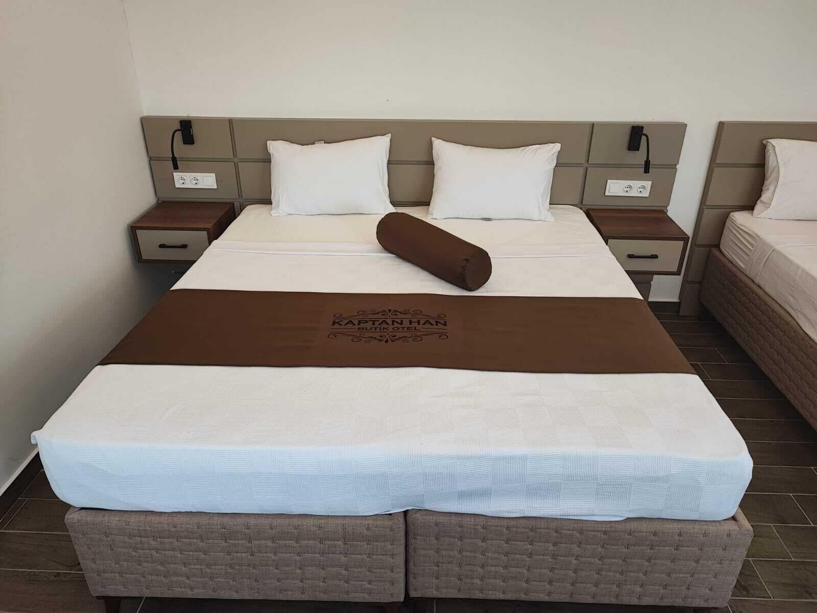 Ein Doppelbett mit brauner Kopfteil, weißer Bettwäsche und einem braunen Kopfkissen. Zwei Nachttische mit Lampen und eine braune Sitzbank am Fußende des Bettes.