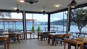 Un espace de restauration intérieur avec des tables en bois et des chaises rembourrées bleues, de grandes fenêtres offrant une vue sur la mer et un éclairage suspendu.
