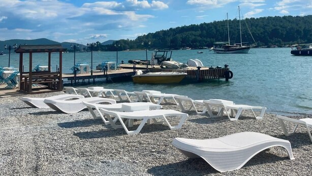 Une plage de galets avec des chaises longues blanches et un kiosque en bois. Des bateaux sont amarrés au loin.