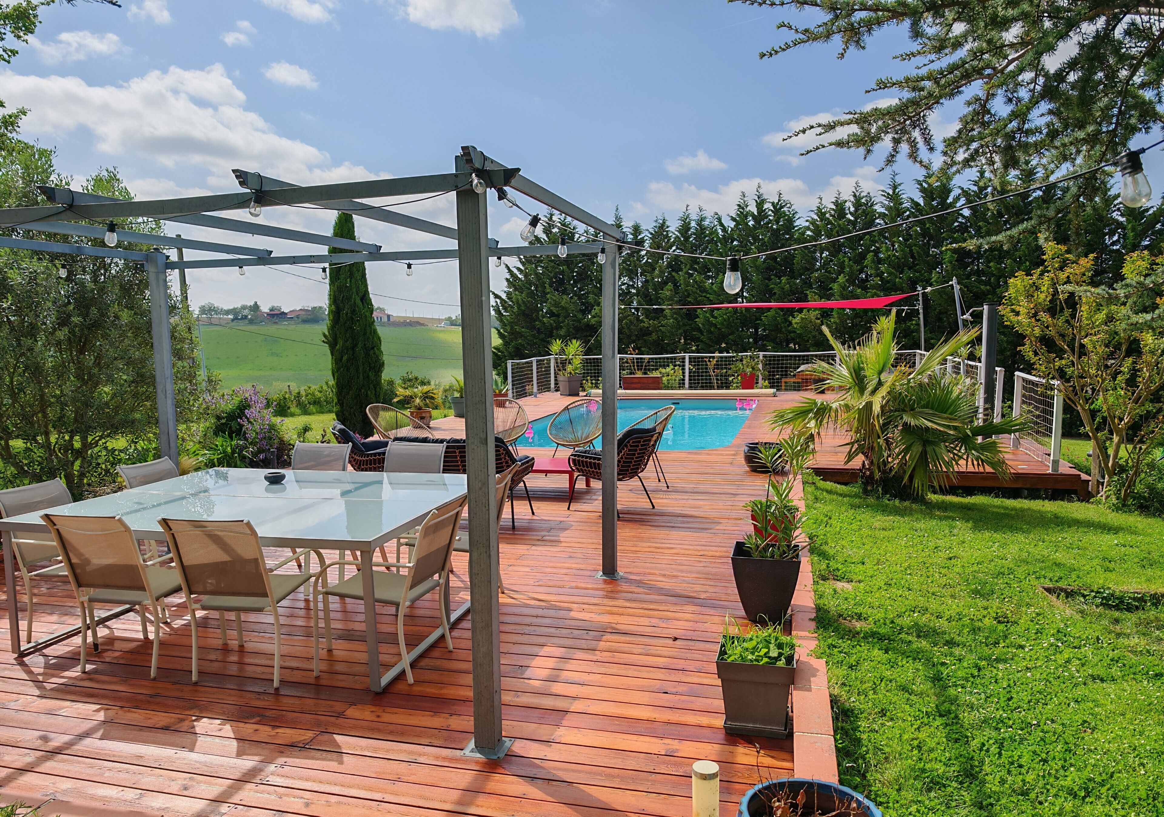 Eine Holzterrasse mit Glastisch und Stühlen, einem Pool und einer Pergola mit Lichterketten.