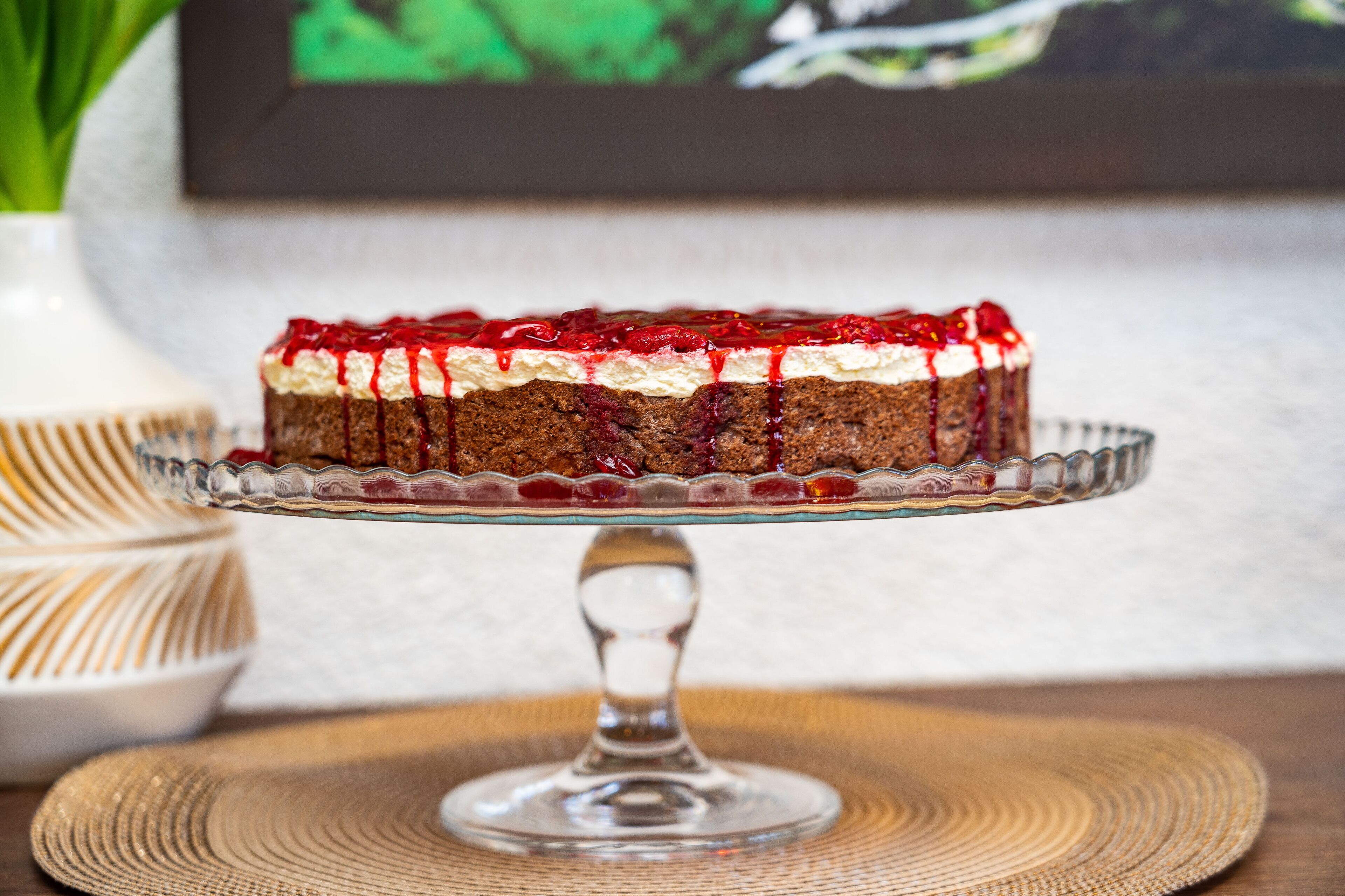 A layered cake with cream and red fruit topping on a glass stand.