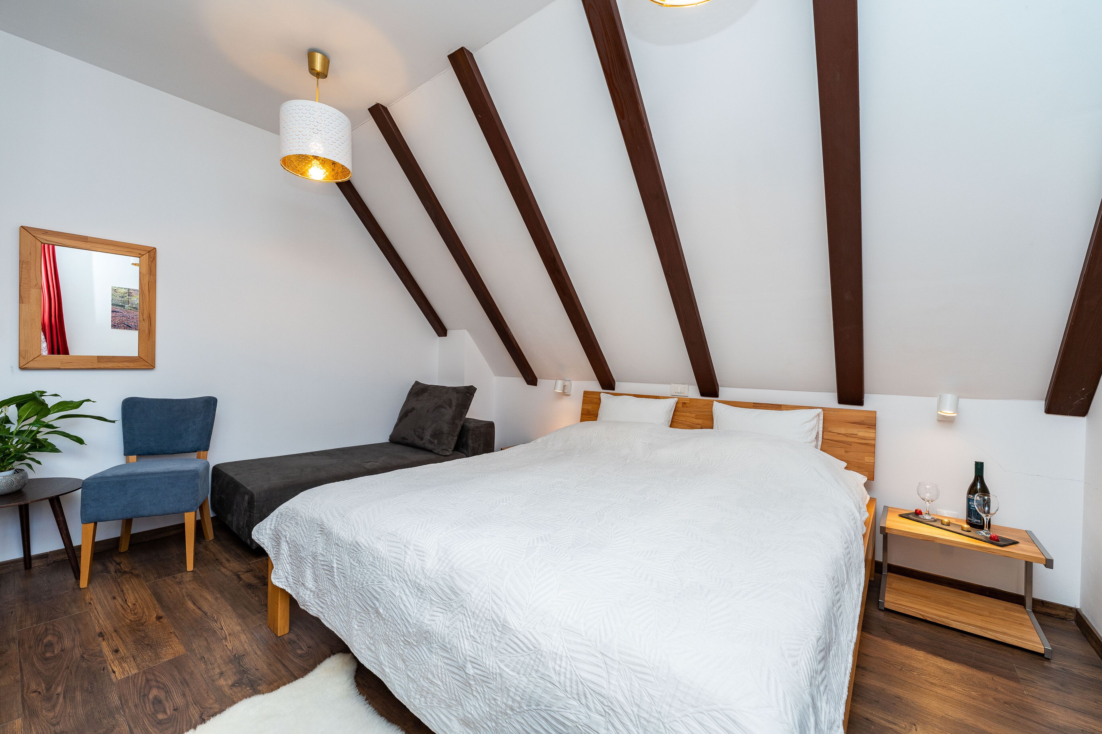 A modern bedroom with a large bed, wooden beams, a desk with a lamp, and a potted plant.