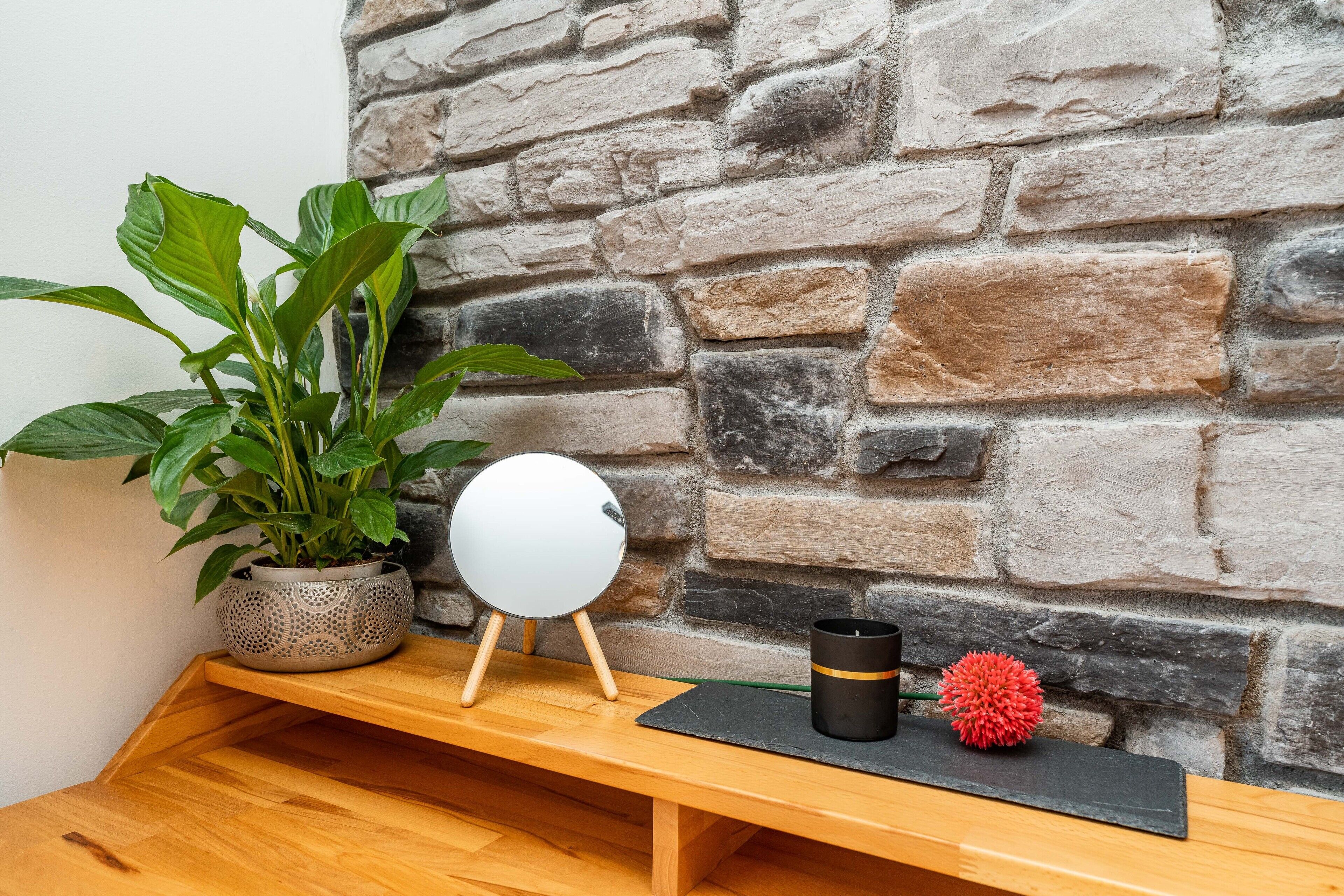 A wooden bench with a potted plant, a round mirror, and a candle holder against a stone wall.