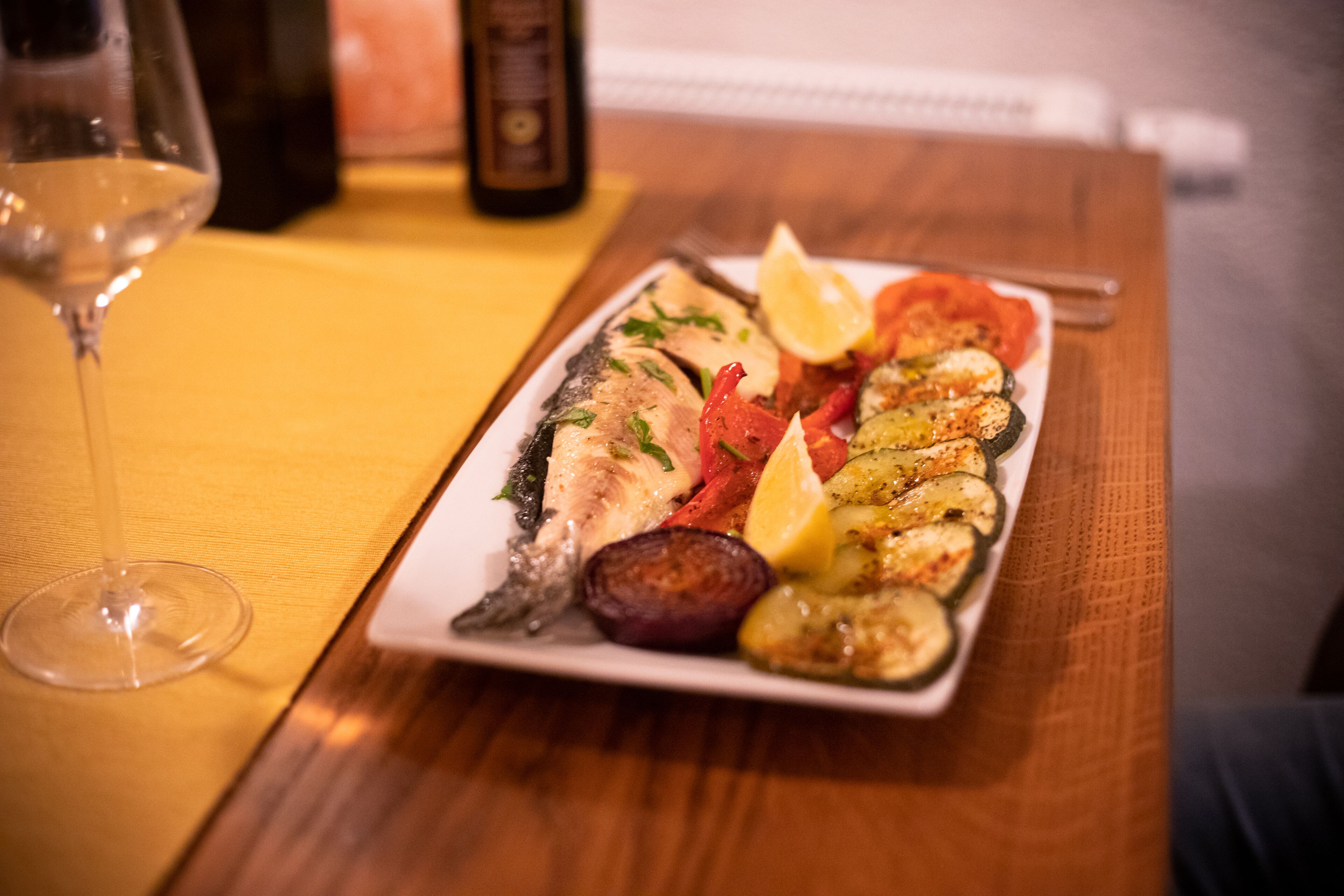A plate of grilled fish and vegetables on a wooden table with a wine glass and a bottle in the background.