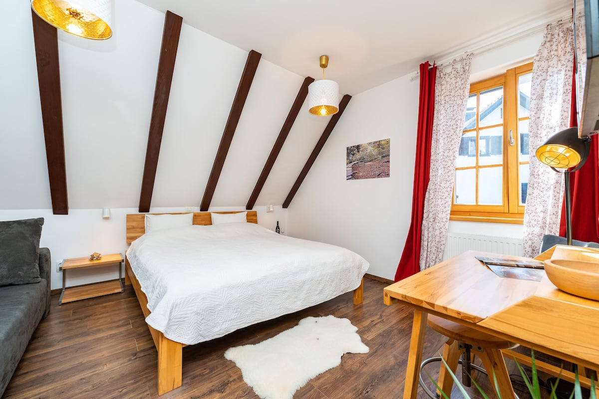 A bedroom with a bed, a wooden table, a small bench, a nightstand, and a window with curtains.
