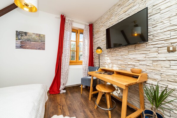 Une pièce avec un bureau en bois, une chaise, un téléviseur fixé au mur en pierre, une fenêtre avec des rideaux rouges et une plante.