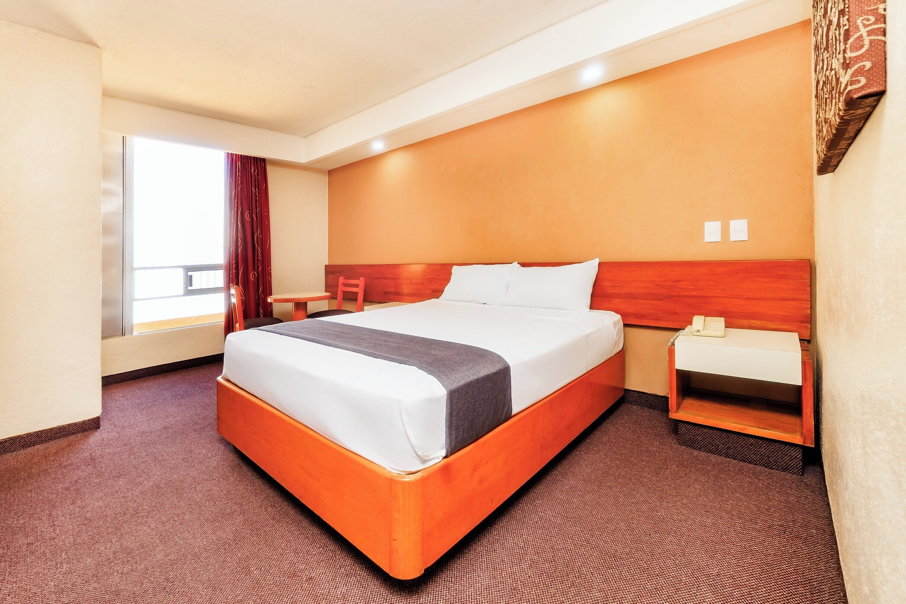 Una habitación de hotel con una cama grande, una mesita de noche, cortinas en una ventana y una obra de arte colgada en la pared.
