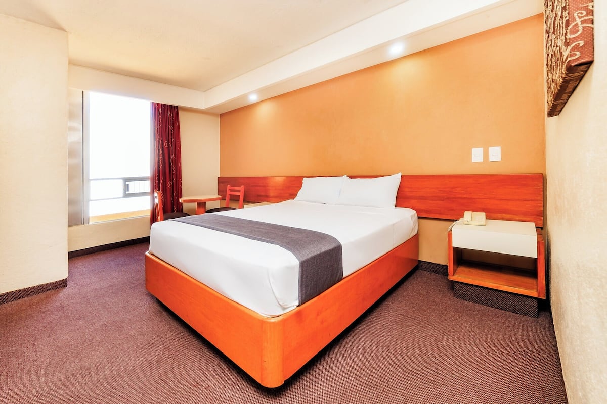Una habitación de hotel con una cama grande, una mesita de noche, cortinas en una ventana y una obra de arte colgada en la pared.