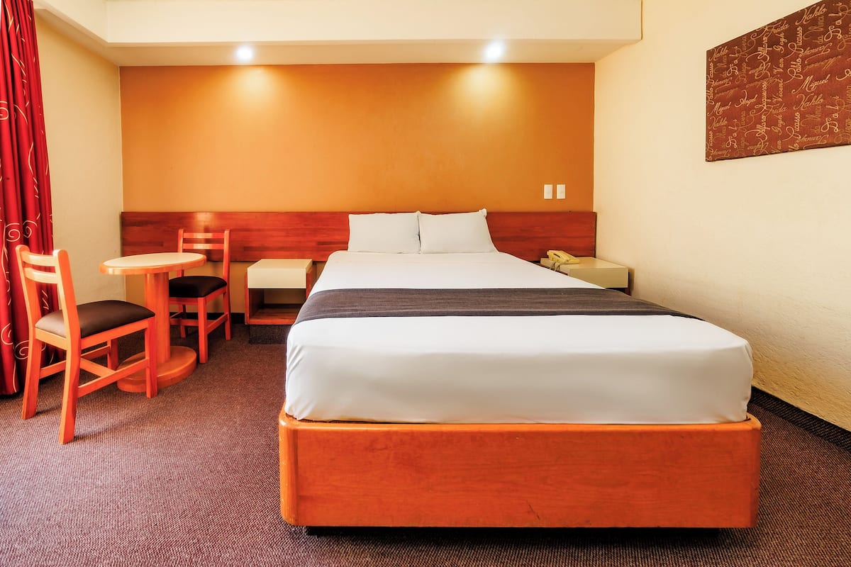 Habitación de hotel con una cama grande, cabecera de madera, una mesita redonda pequeña, dos sillas, un escritorio con teléfono y una cortina roja.