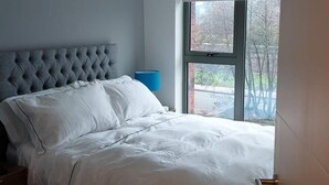 Ein Schlafzimmer mit einem gebürsteten Kopfteil, einem Bett mit weißer Bettwäsche, einem Nachttisch, einer blauen Lampe und einem Fenster mit Blick auf Bäume und eine Straße.