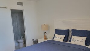 Ein Schlafzimmer mit Bett, Deckenventilator, Nachttisch mit Lampe und einem durchgegangenen Türspalte sichtbaren Badezimmer.