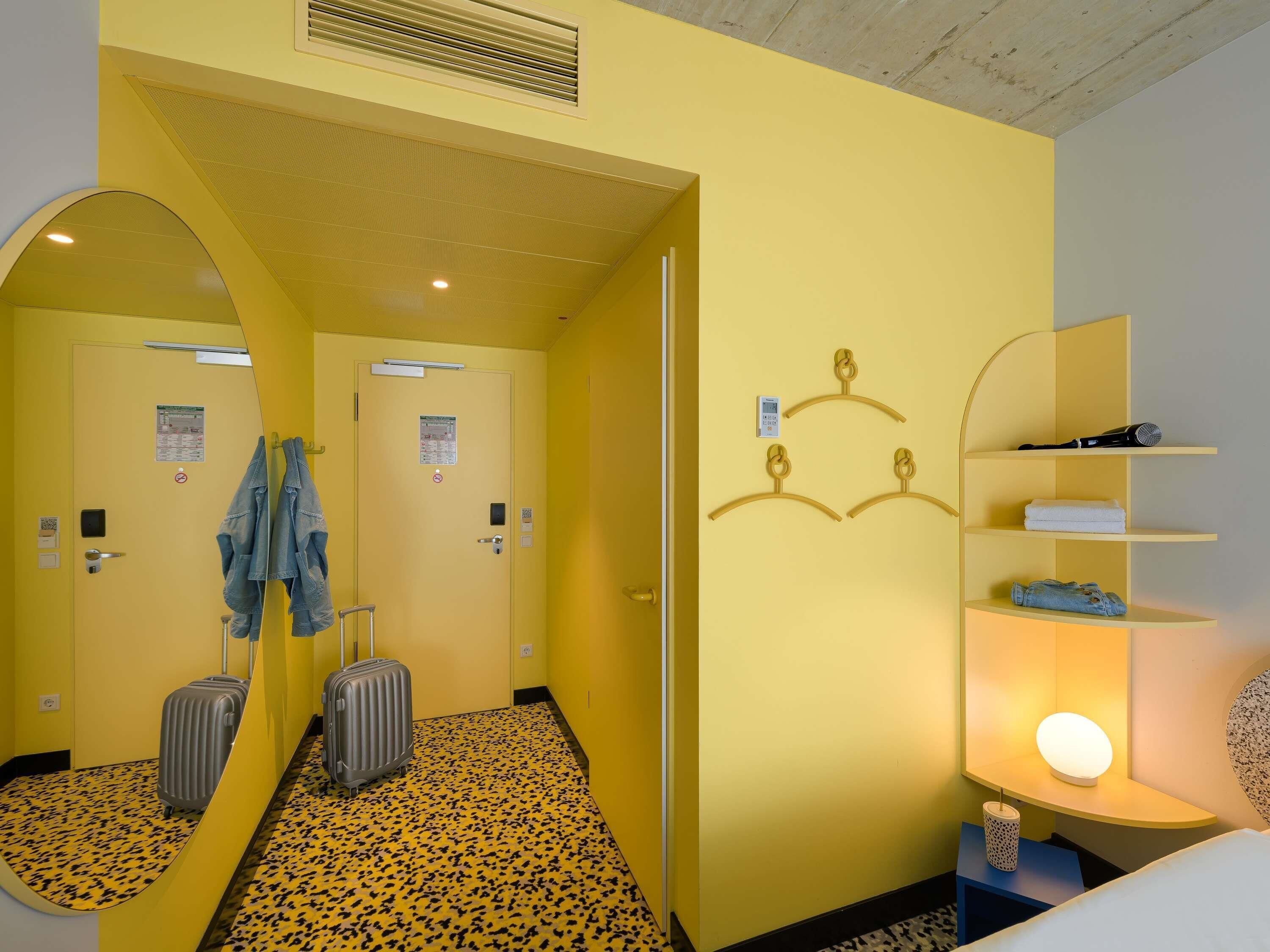 Habitación de hotel con pared espejada, una cama, mesita de noche con lámpara, armario y maleta.