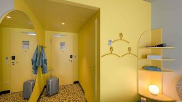 Habitación de hotel con pared espejada, una cama, mesita de noche con lámpara, armario y maleta.