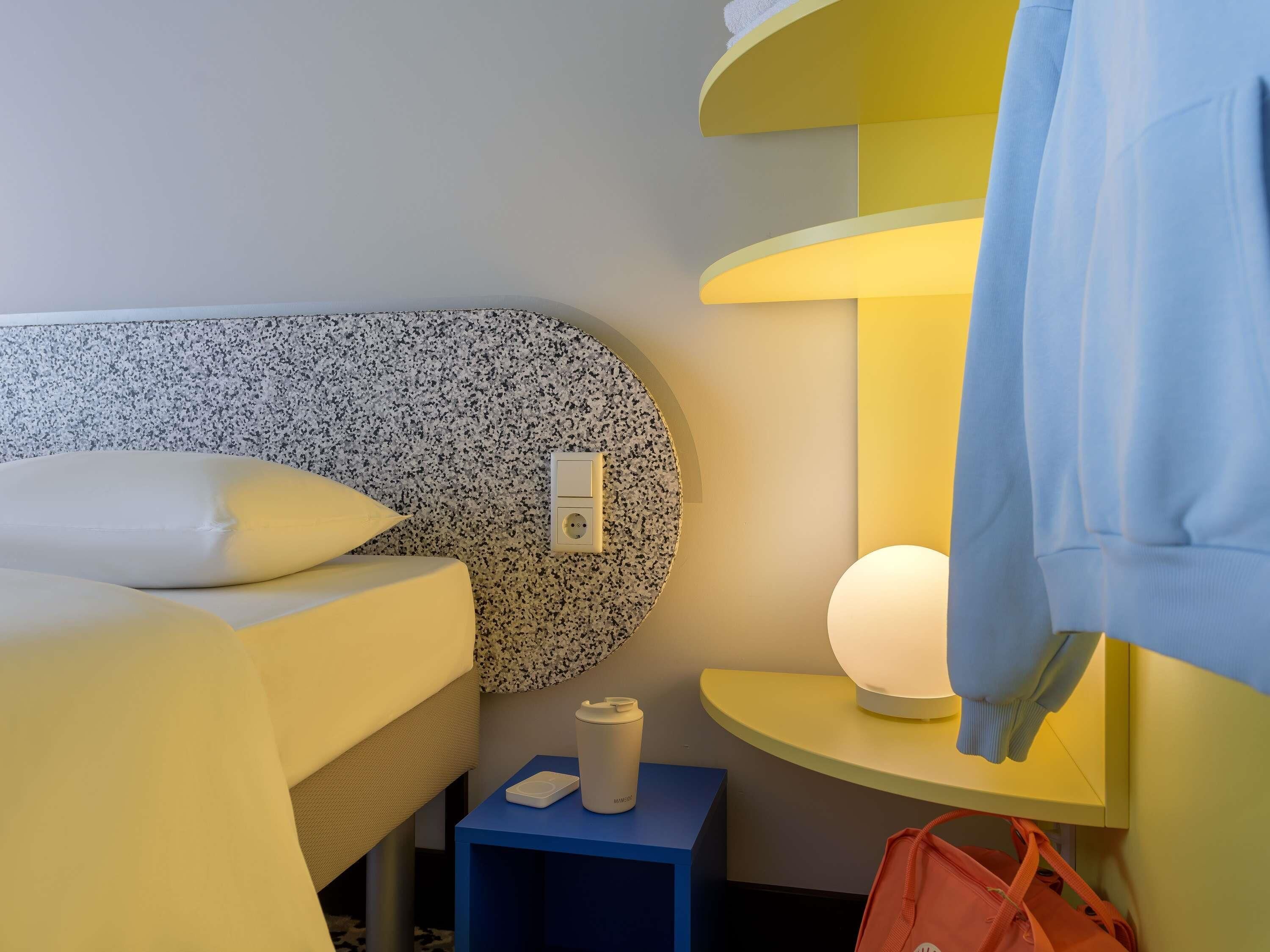 Habitación de hotel con cama, mesita de noche con lámpara, enchufe y un pequeño tocador azul.
