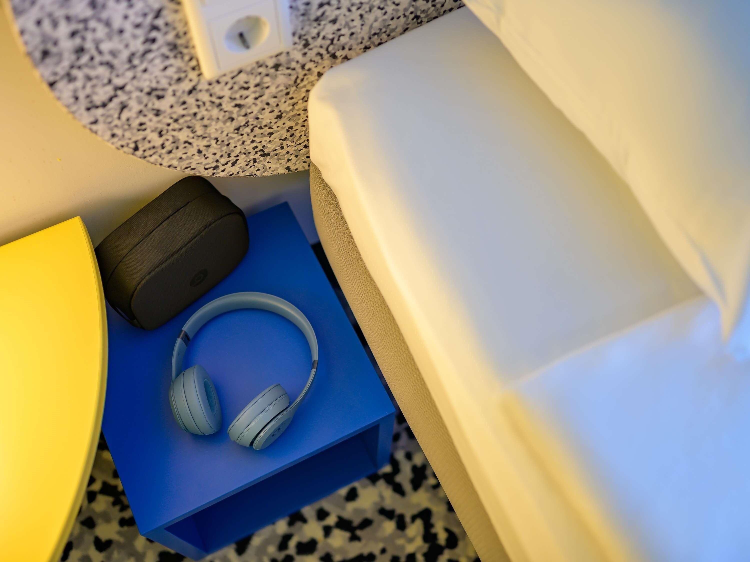 Una cama con un almohadón blanco, una mesita de noche azul con auriculares y un estuche de carga negro.