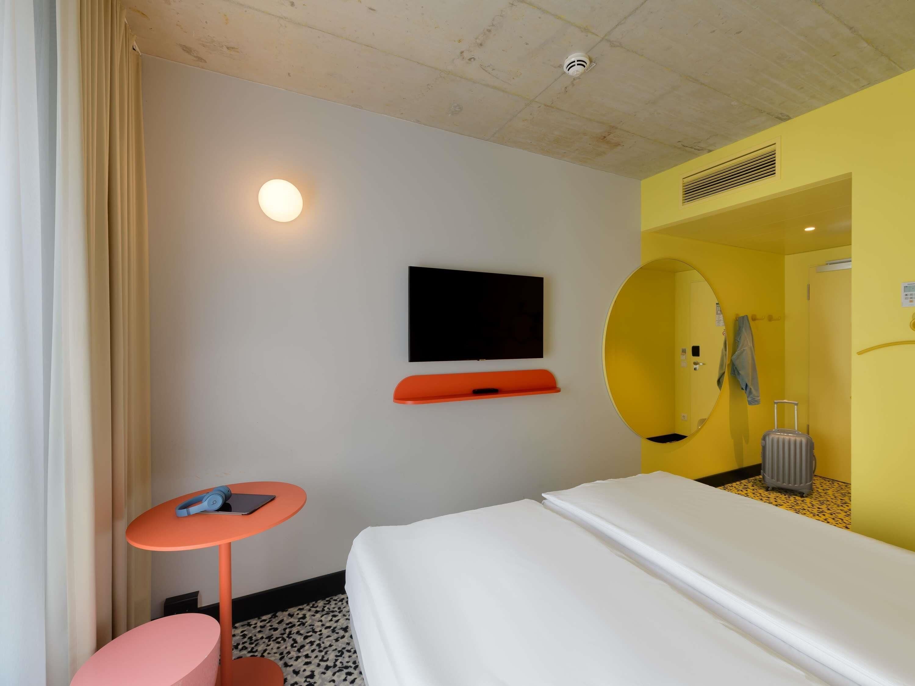 A hotel room with a bed, a small table, a TV, a mirror, and a suitcase.
