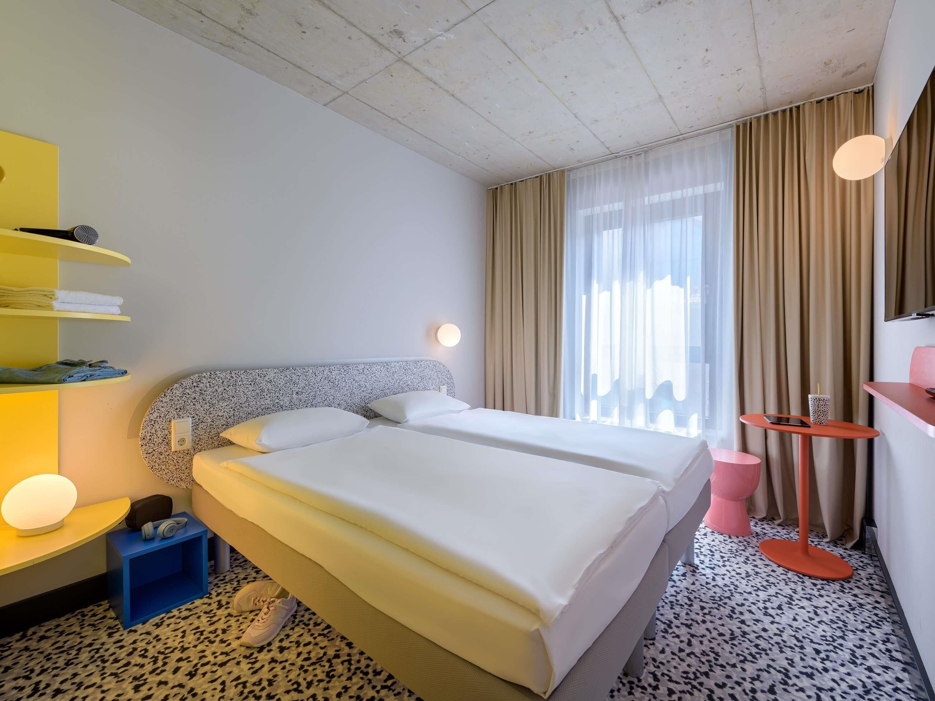 A hotel room with a double bed, a small desk with a chair, a shelf with towels, and a large window with curtains.