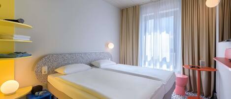 Quarto de hotel com uma cama de casal, uma pequena escrivaninha com cadeira, um armário com toalhas e uma grande janela com cortinas.