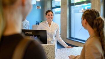 A reception desk with a person standing behind it, a computer, and a window with a view outside.
