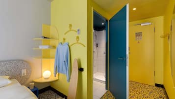Habitación de hotel con una cama, una mesita de noche, una lámpara, una toalla azul y una puerta de madera que da a un baño.