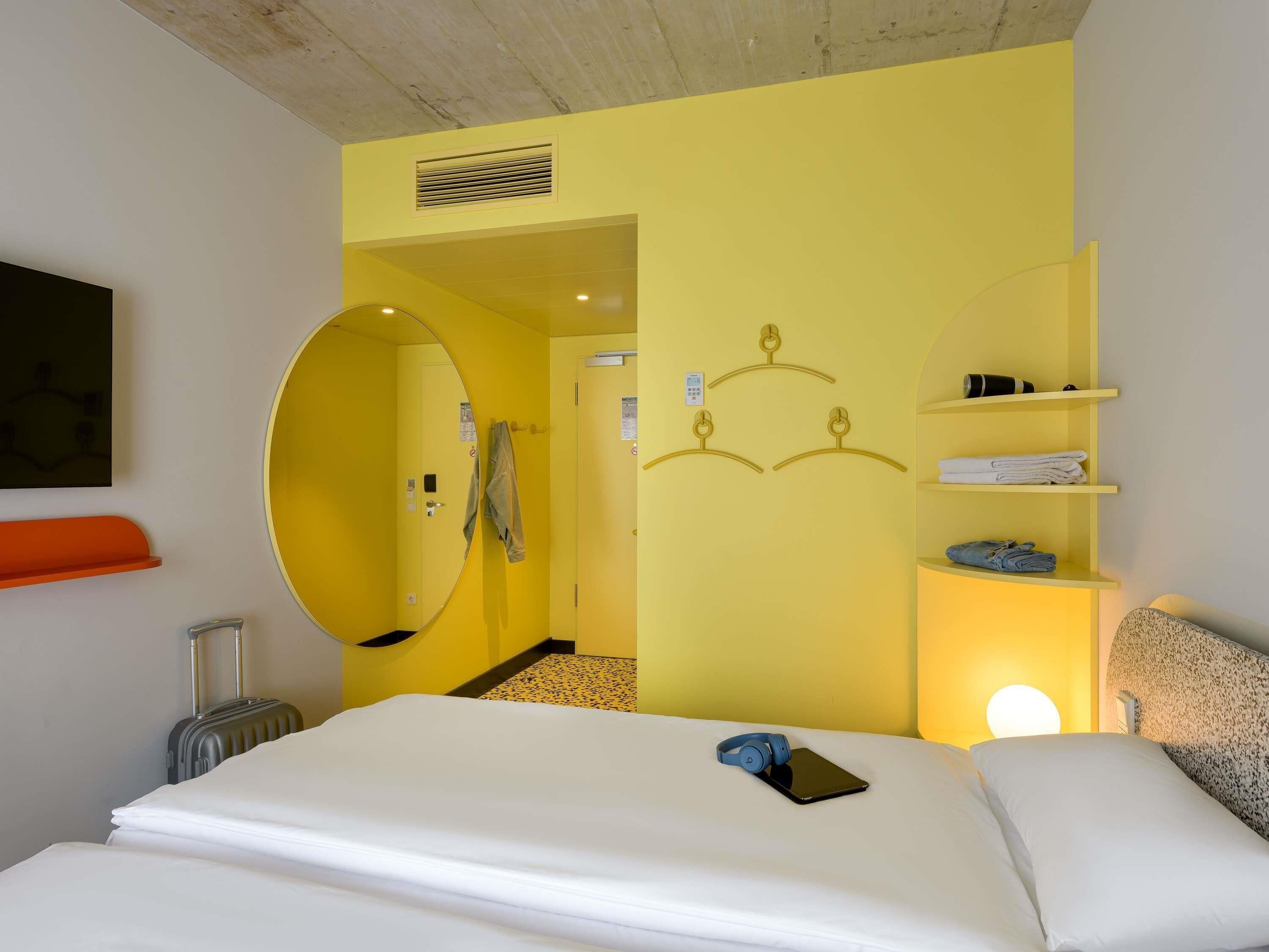 A hotel room with a bed, a mirror, a shelf with towels, a suitcase, and a wall-mounted light.