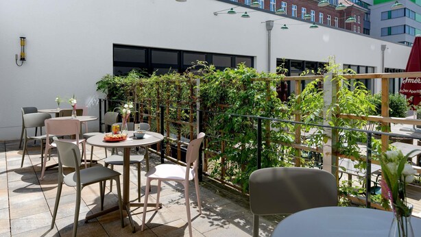 Een terras buiten met ronde tafels en stoelen, een houten treillis met klimplanten, en een gebouw met grote ramen op de achtergrond.