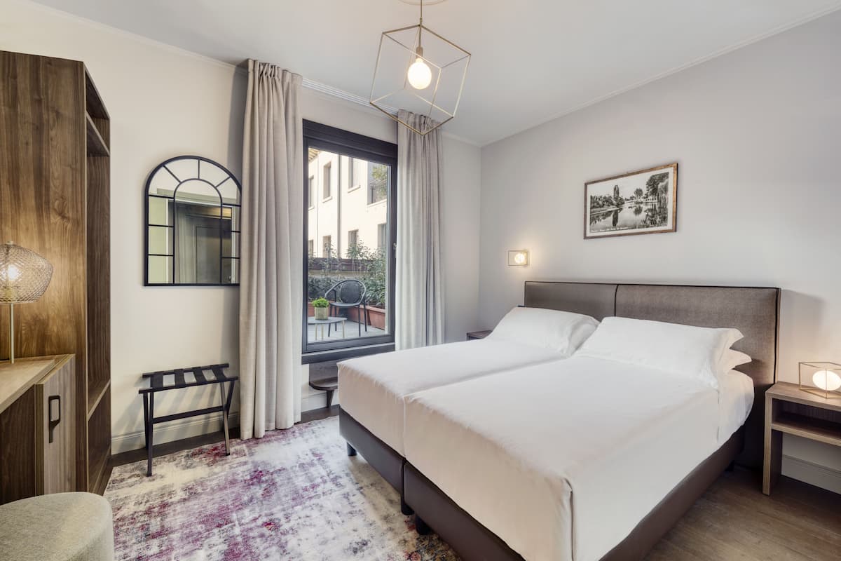 A modern bedroom with a large bed, a wooden wardrobe, a bench, a window with curtains, and a framed picture on the wall.
