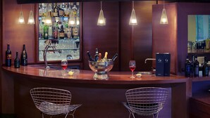 Uma área de bar com duas cadeiras vazias, balcão com garrafas e copos, e luminárias pendentes.