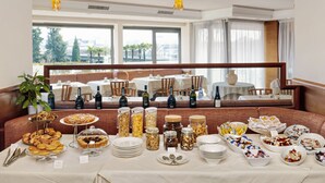 Disposição de buffet com diversos alimentos e acompanhamentos, área de refeições com mesas e cadeiras, e grandes janelas com vista para a cidade.