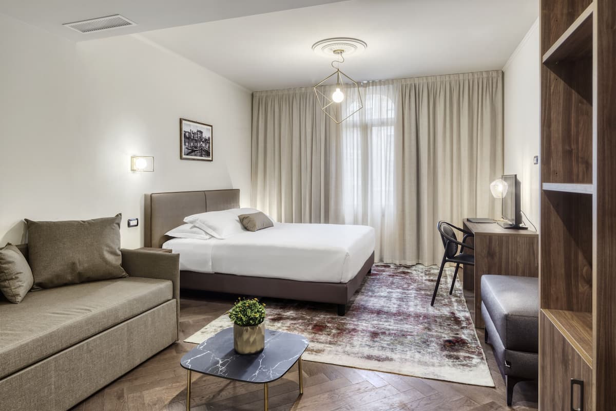 A modern hotel room with a bed, sofa, desk, and a small table with a plant.