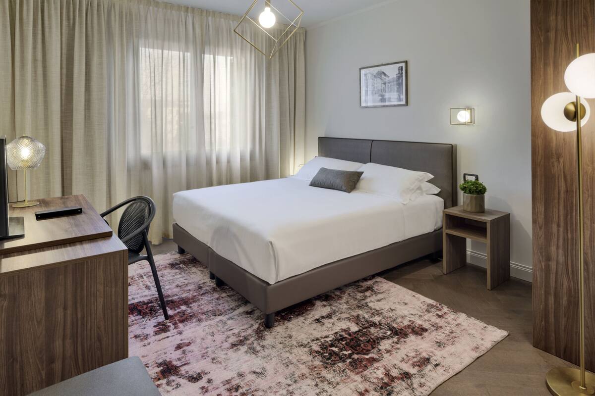 A modern hotel room with a large bed, a desk, a chair, a nightstand, and a floor lamp.
