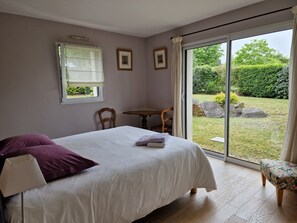 Quarto com cama, uma cadeira, uma mesinha e vista para um jardim através de uma porta de correr.