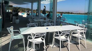 Un espace de restauration en plein air, avec des chaises et des tables blanches, offrant une vue sur une mer d’un bleu profond et sur une côte lointaine.