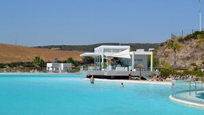 Un espace moderne aménagé au bord de la piscine, avec un bâtiment blanc portant l’inscription « ALCAZABA », des chaises longues et une piscine à l’eau d’un bleu limpide.