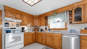 A kitchen with wooden cabinets, a stove, a dishwasher, and a window with a view. - Charming Cottage Steps from Hulls Cove Beach & Entrance to Acadia! (Bar Harbor)