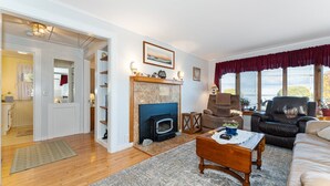 A living room with a fireplace, a sofa, a coffee table, and a view of the outdoors. - Charming Cottage Steps from Hulls Cove Beach & Entrance to Acadia! (Bar Harbor)