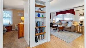 A living room with a bookshelf, a sofa, and a coffee table. - Charming Cottage Steps from Hulls Cove Beach & Entrance to Acadia! (Bar Harbor)