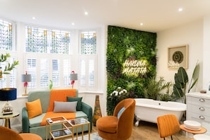 A modern living room with a green sofa, orange armchairs, a glass coffee table, and a large window with stained glass.