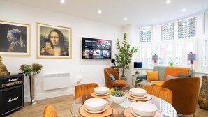 Un salón moderno con una mesa de vidrio dispuesta para cuatro personas, rodeada de sillas naranjas. Un televisor de pantalla plana montado en la pared muestra un servicio de streaming.