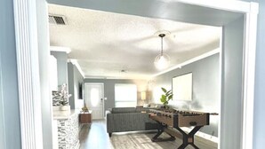 A spacious living room with a wooden floor, a grey sofa, a coffee table, and a foosball table. - Spacious & Luxurious Villa - Large Fenced Backyard (West Palm Beach)