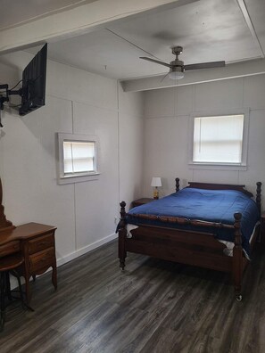 A bedroom with a wooden bed, a ceiling fan, a TV mounted on the wall, and two windows with blinds.