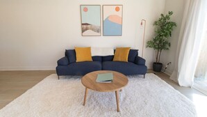 Un salon avec un canapé bleu foncé, deux coussins jaunes, une table basse ronde en bois et un tapis blanc à longues mèches.