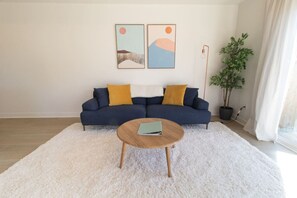Un salon avec un canapé bleu foncé, deux coussins jaunes, un tapis blanc et moelleux, une table basse ronde en bois sur laquelle repose un livre, et deux tableaux abstraits accrochés au mur.